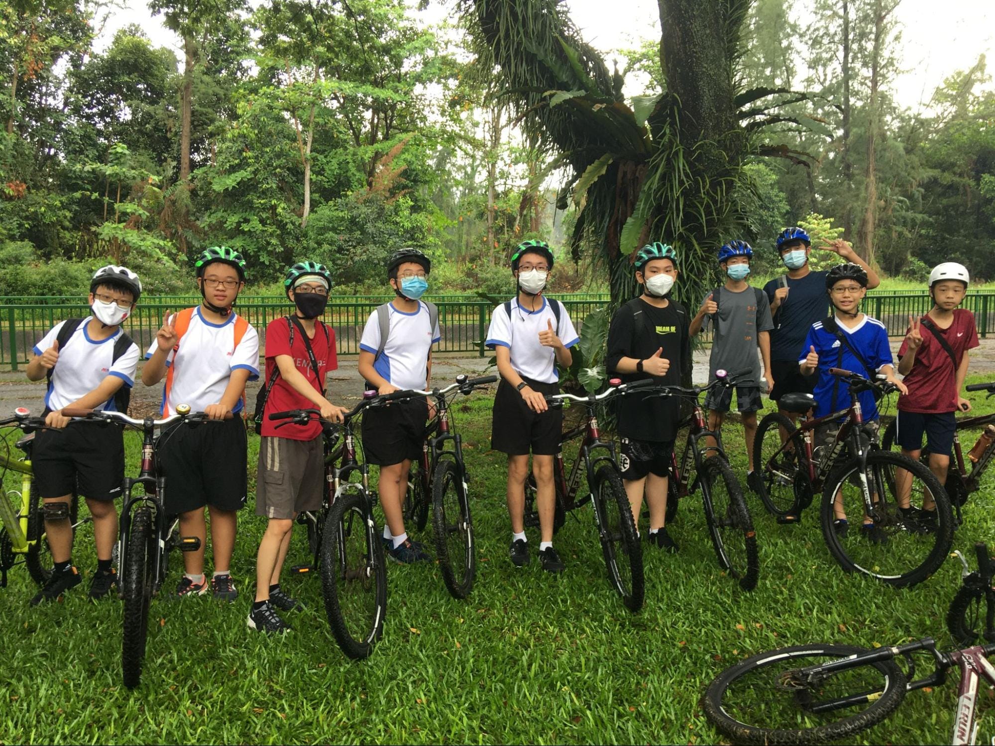 Ten young cyclists wearing helmets and face masks pose with bicycles on a grassy park path surrounded by trees.